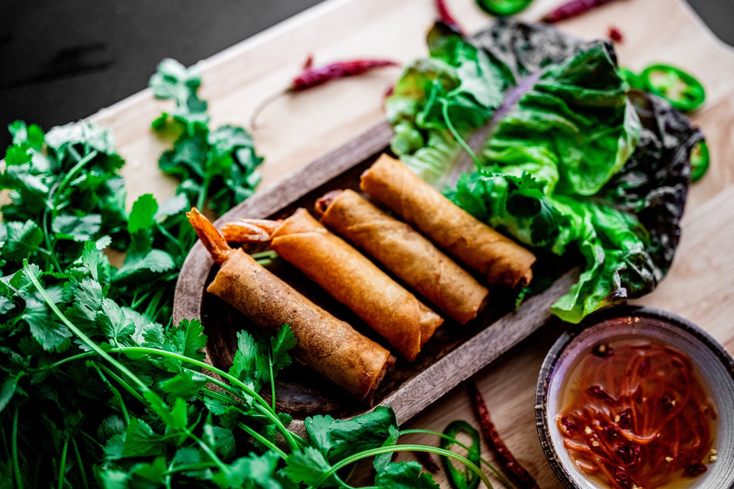 Crispy Vietnamese egg rolls on a wooden board with fresh cilantro, lettuce and chili dipping sauce